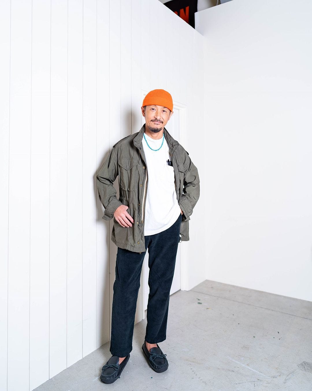 Man standing in corner wearing orange hat, white shirt, olive jacket, turquoise necklace, dark pants and loafers, men's autumn date outfit