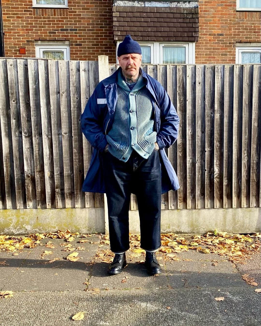 man standing on the sidewalk in front of a fence, wearing a long blue coat, light blue cardigan, dark blue jeans, and black boots