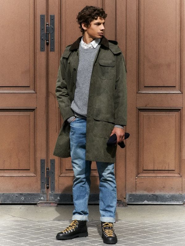 man standing in front of large doors wearing a long waxed jacket with a grey sweater, blue jeans, and black boots