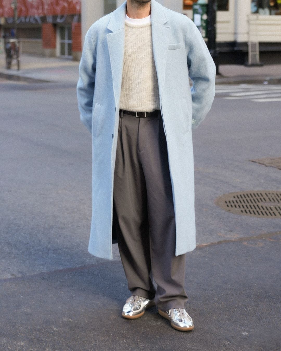 close-up of a man wearing a long light blue topcoat, grey sweater and grey dress pants