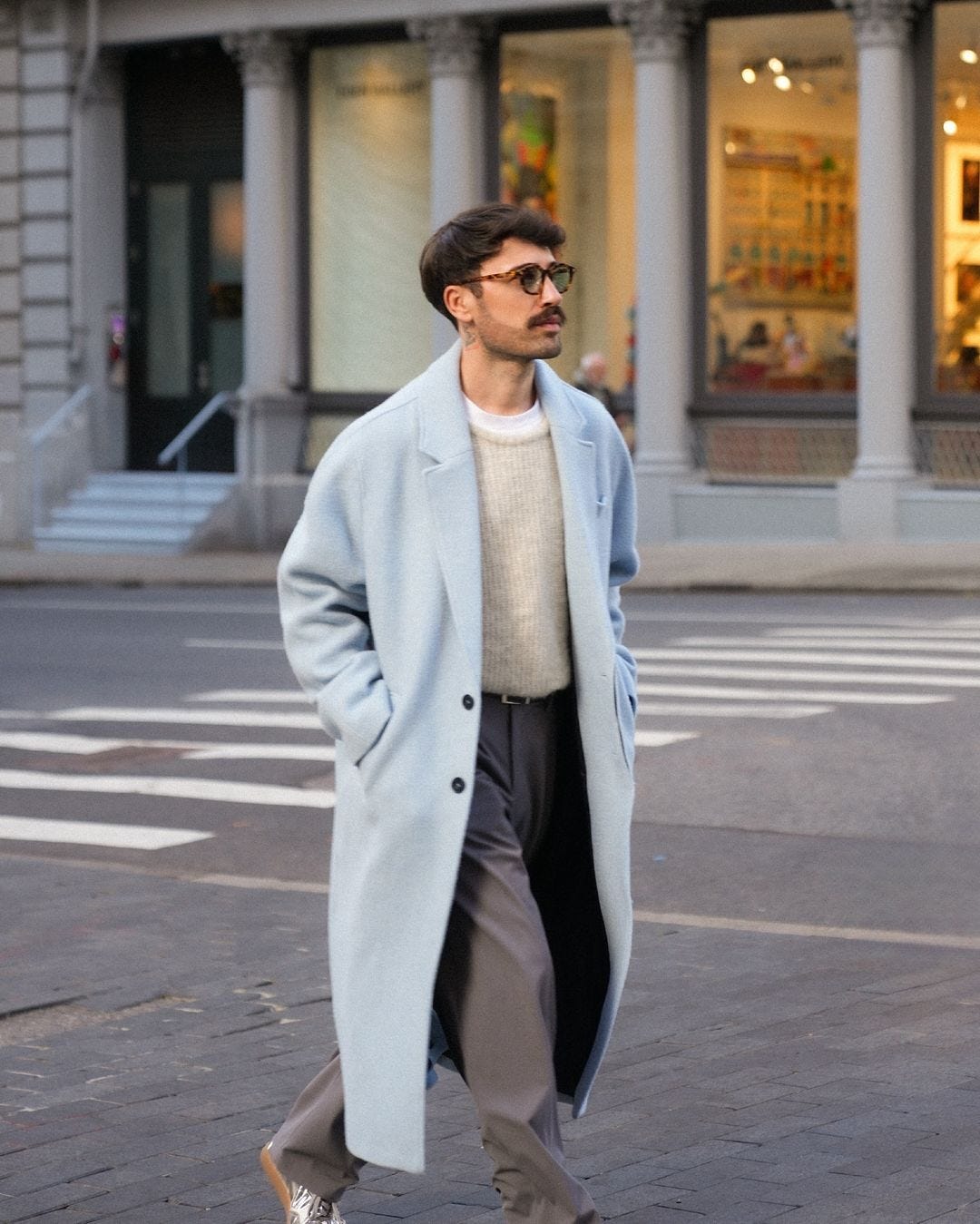 man walking down a city street wearing a long light blue topcoat, grey crewneck sweater, and grey trousers with black belt