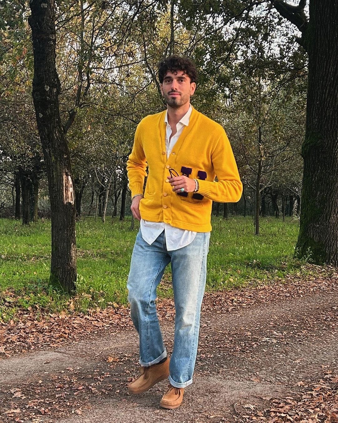 stylish man wearing a yellow letterman cardigan over a white oxford shirt with light wash jeans and brown Wallabee boots, outfit idea for what to wear on thanksgiving