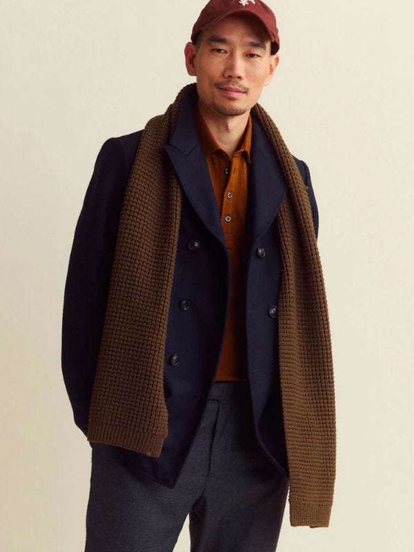 man from the waist up wearing a navy peacoat over an orange knit polo with a brown textured scarf