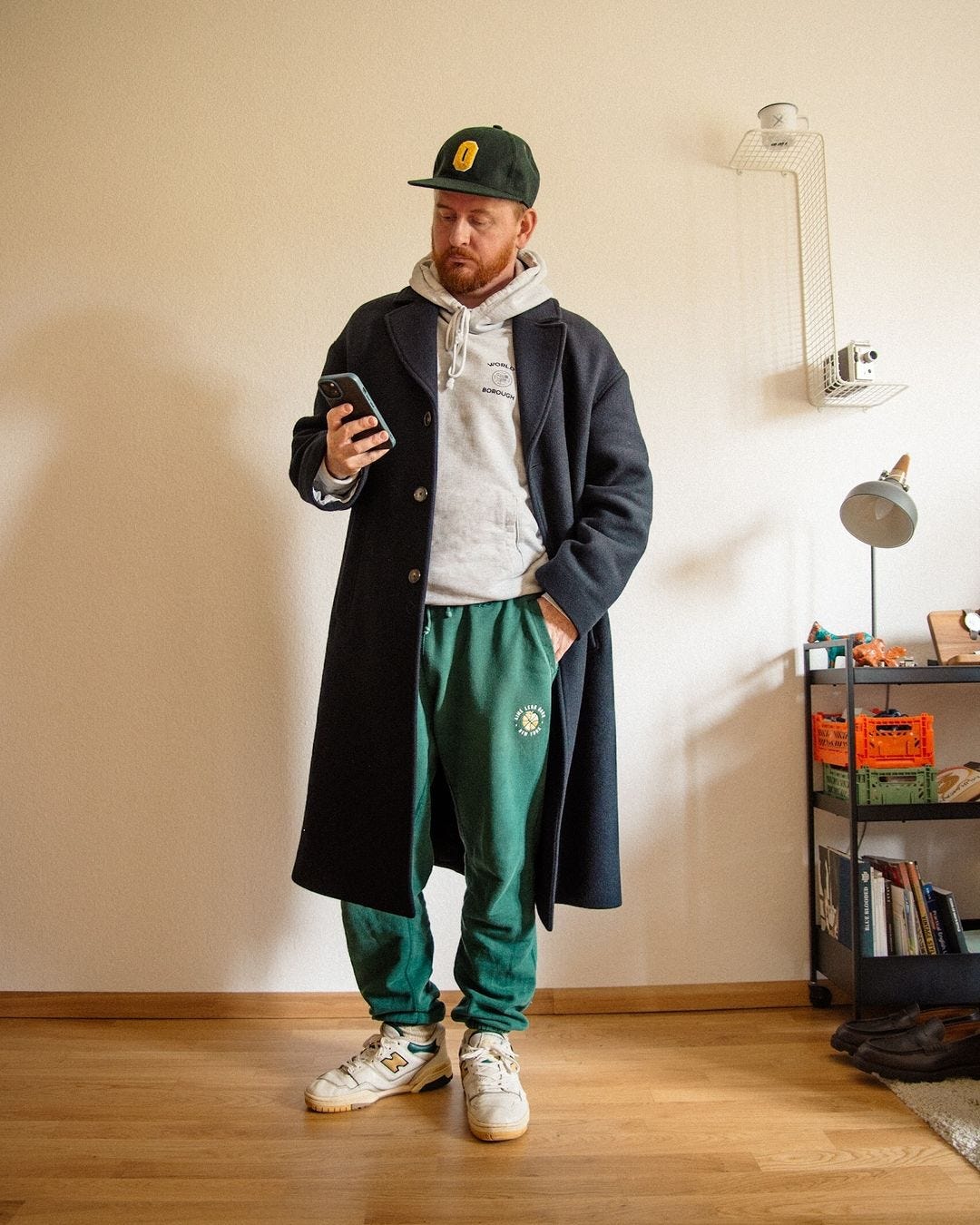 man looking at his phone wearing a navy topcoat, grey hoodie sweatshirt, green sweatpants, and New Balance sneakers