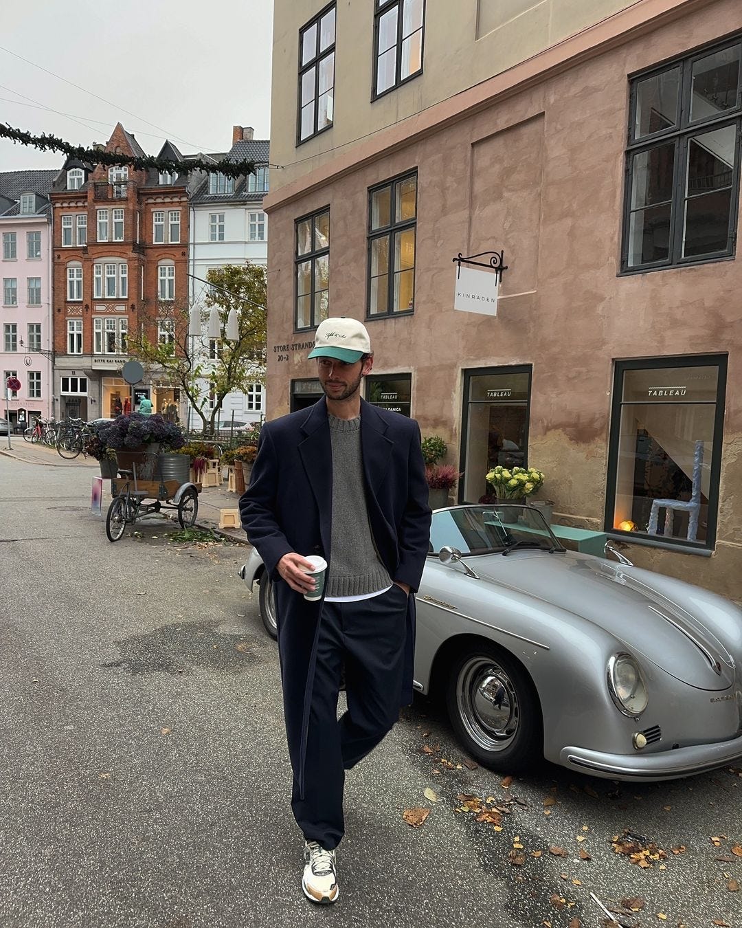 man walking down a city street in front of a silver convertible wearing a navy long coat and holding a cup of coffee