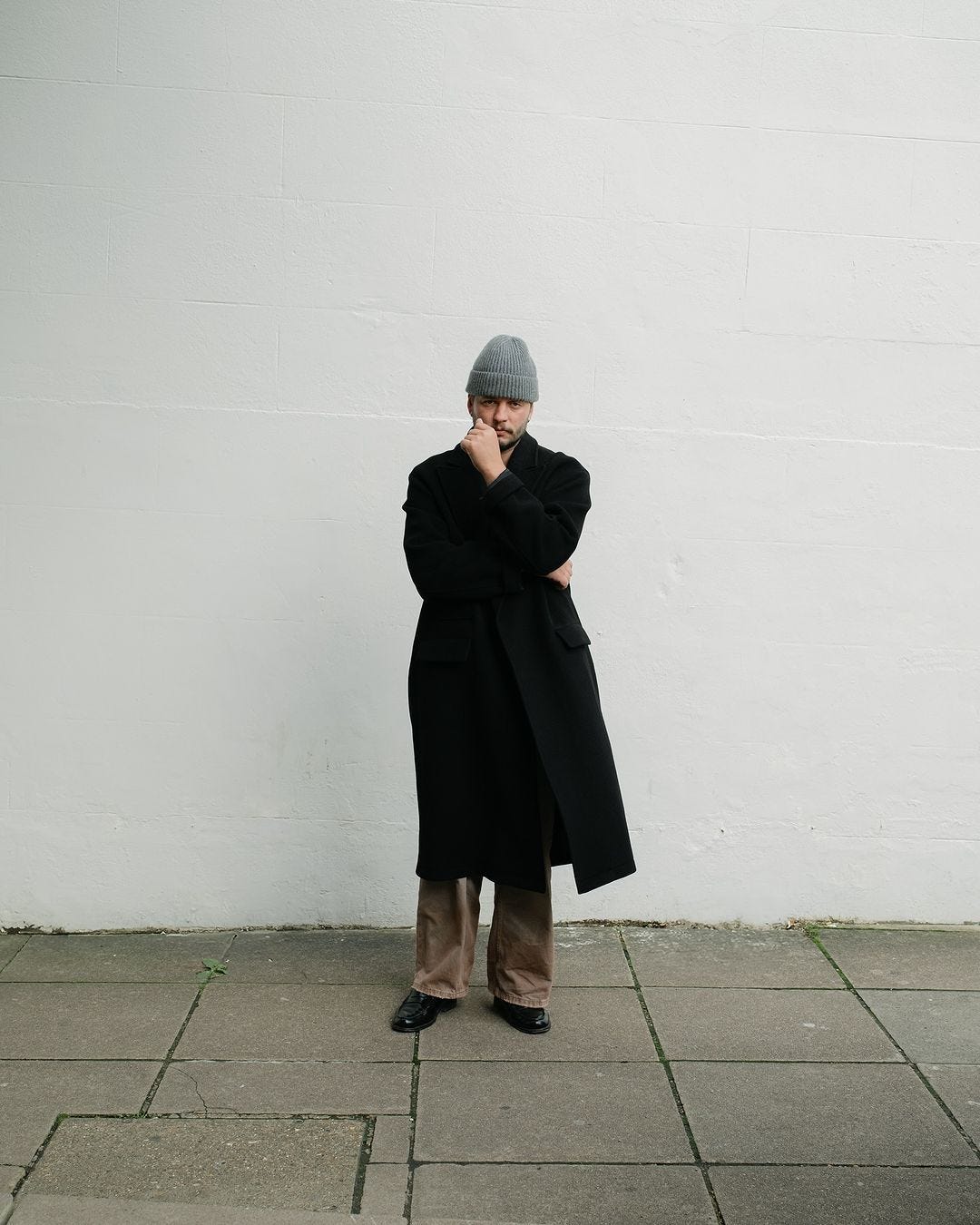 stylish man standing in front of a white wall wearing a grey beanie, black topcoat, brown pants, and black shoes