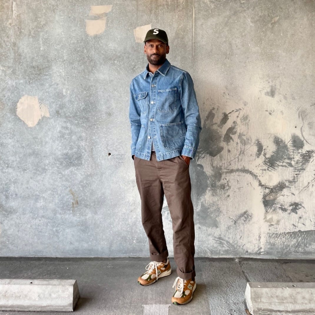 stylish man wearing a baseball cap, blue chore coat, brown pants, and brown New Balance sneakers