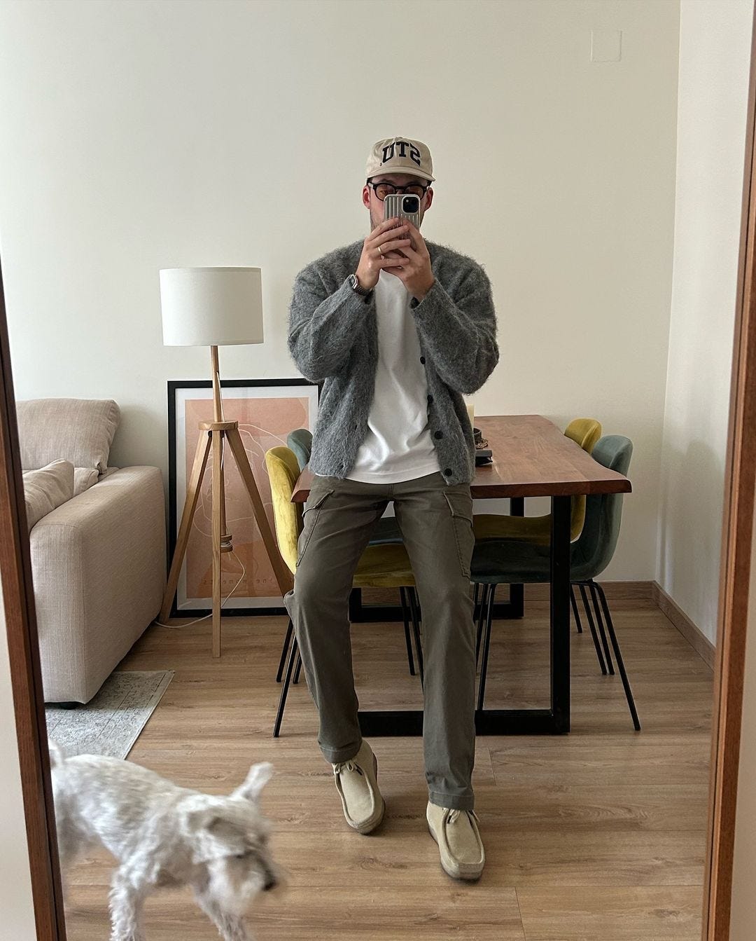 man leaning against a table, taking a selfie in the mirror, wearing a grey cardigan, white t-shirt and olive green pants