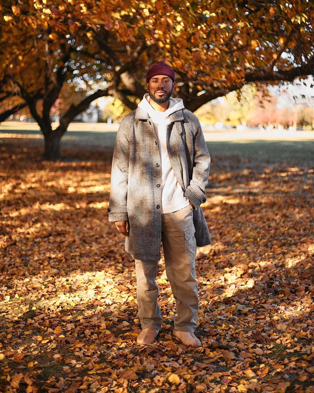 man standing outside in front of a tree with leaves on the ground, wearing a maroon beanie, patterned topcoat, cream-colored hoodie, khaki pants, and light brown boots