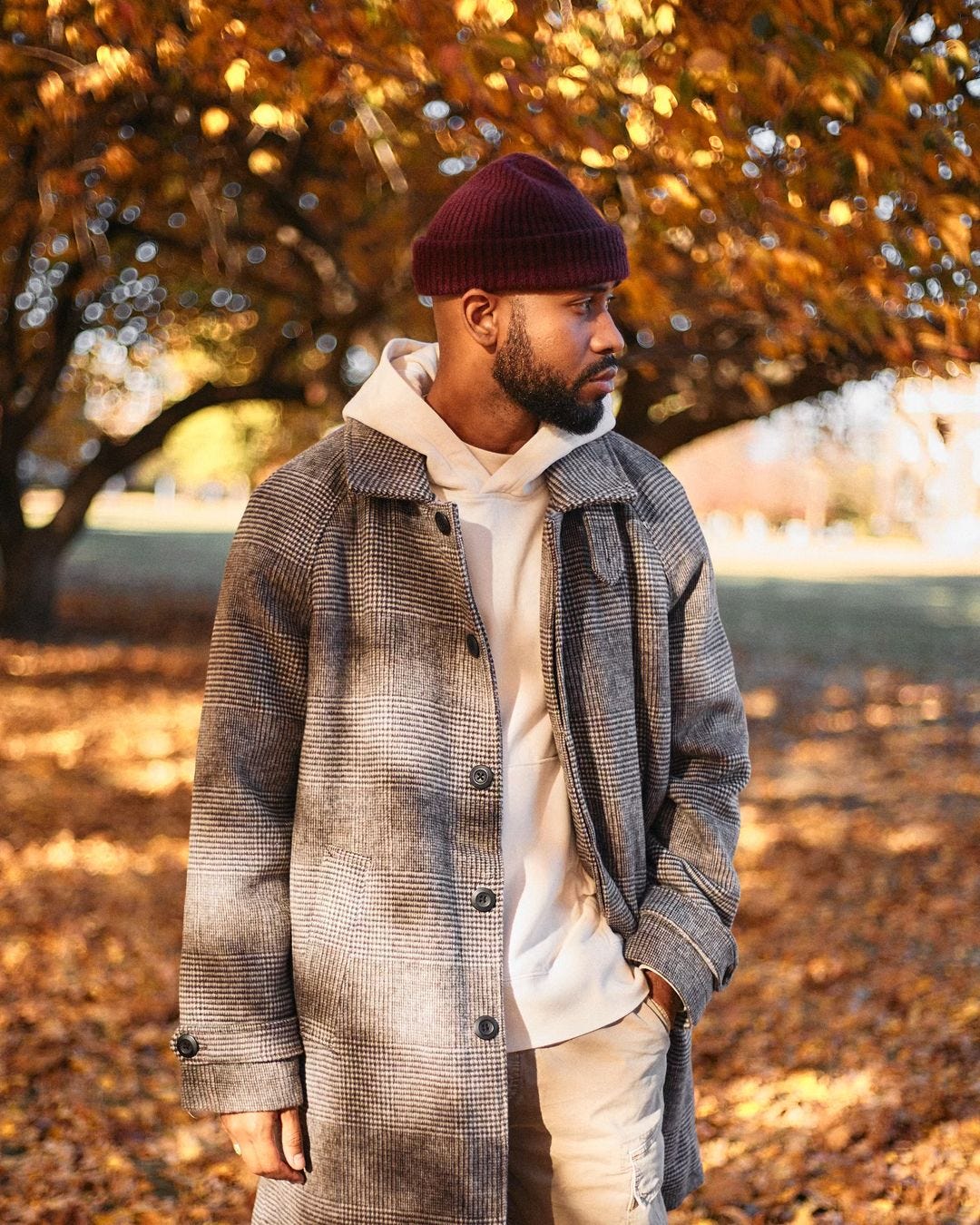 close-up of a man standing outside in front of a tree with leaves on the ground, wearing a maroon beanie, patterned topcoat, cream-colored hoodie, and khaki pants