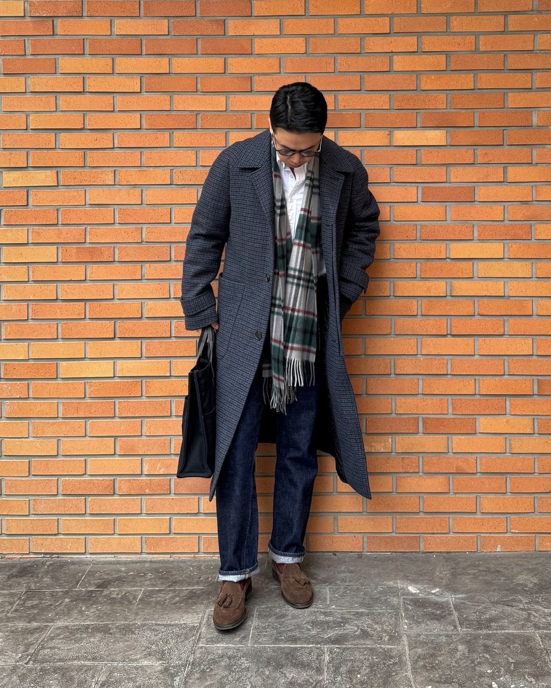 stylish man wearing a patterned topcoat with a plaid scarf, white button down shirt, dark jeans, and brown tassel loafers