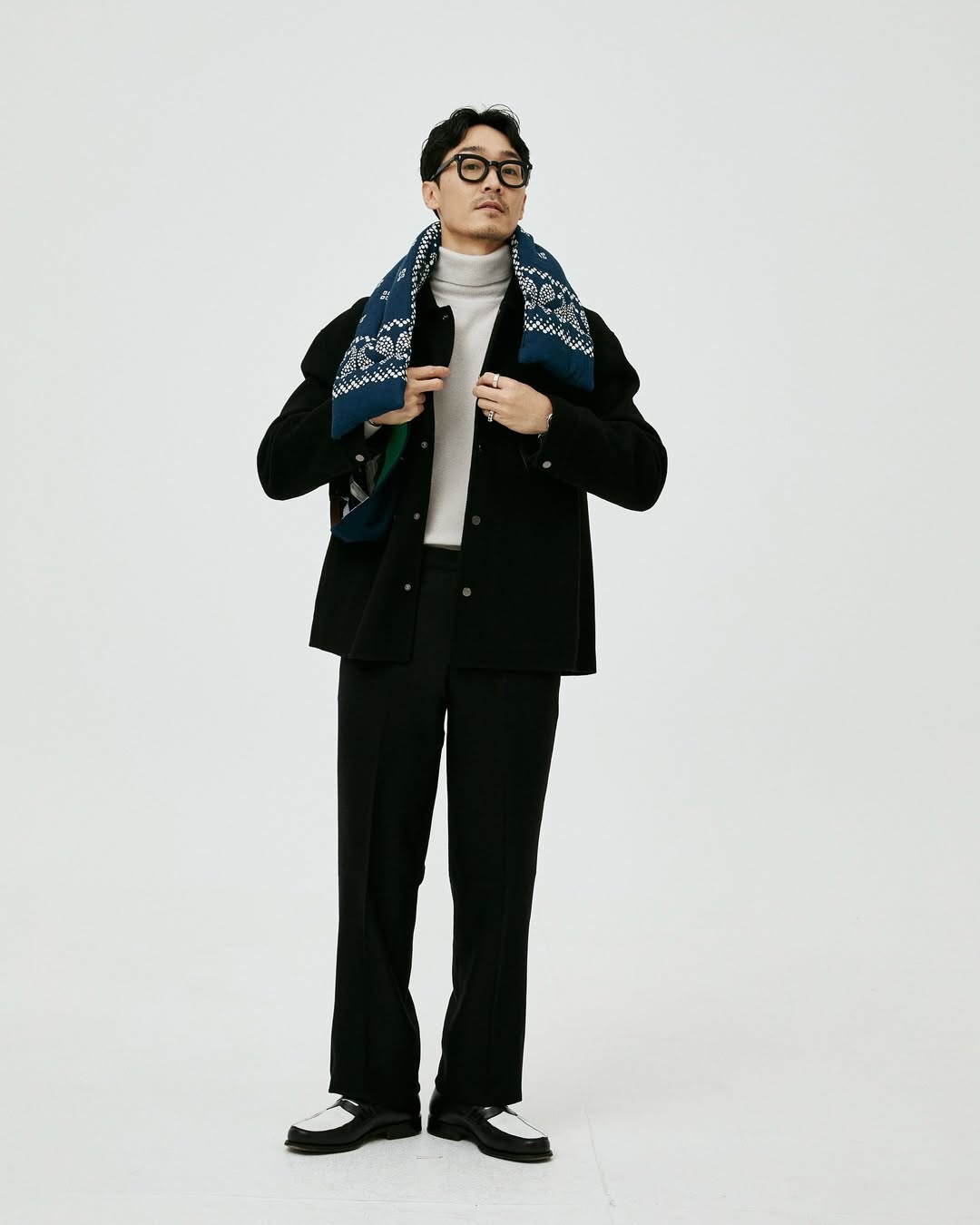 stylish young man in glasses wearing a black jacket and pants with a blue and white quilted scarf hanging around his neck