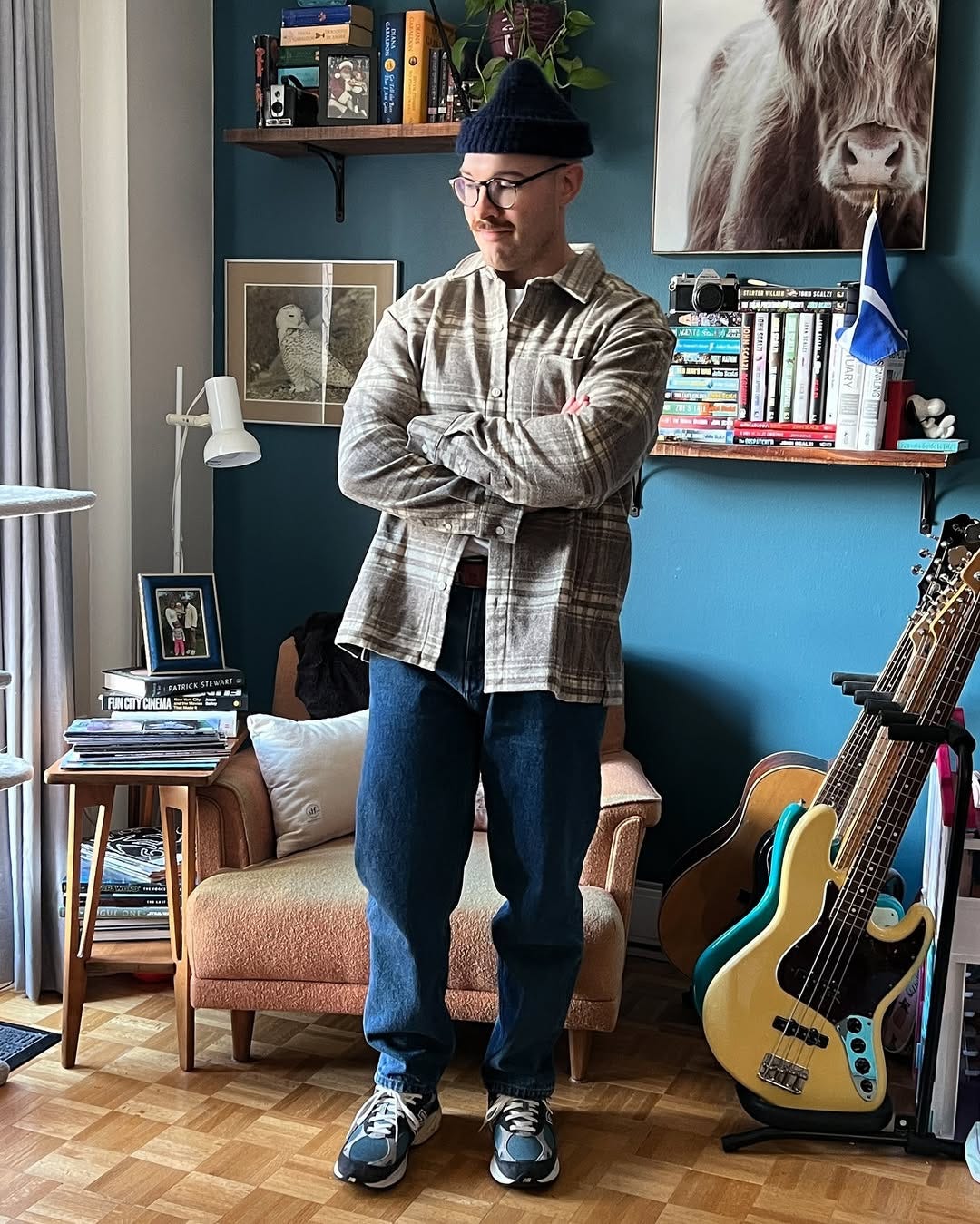 man with his arms crossed wearing a dark beanie, plaid flannel shirt, blue jeans, and blue sneakers