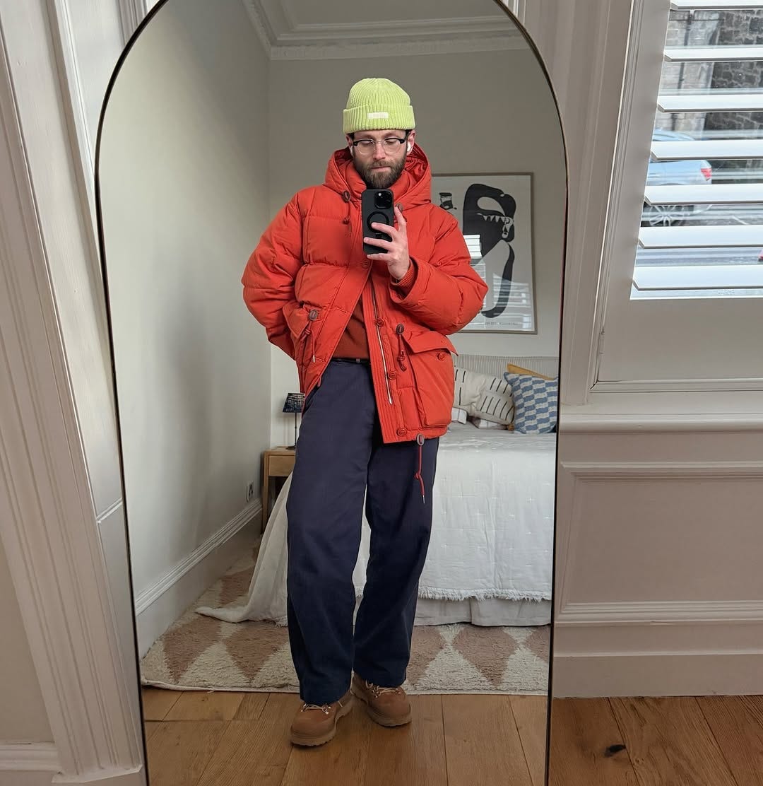 man taking a mirror selfie wearing a green beanie, red puffer jacket, orange sweater, navy pants, and brown boots