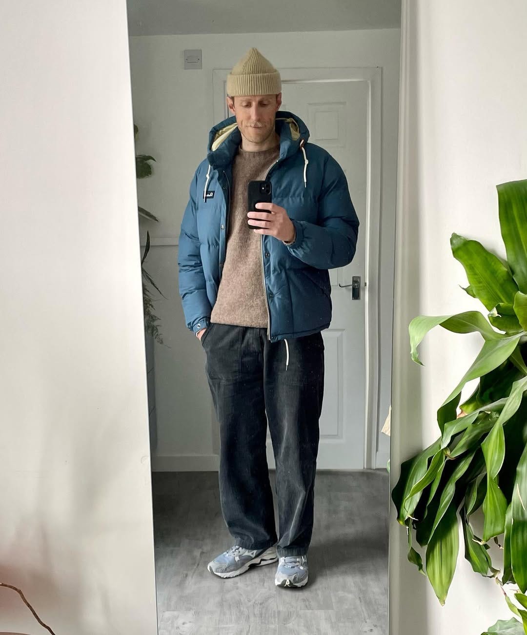 man taking a mirror selfie wearing a tan beanie and sweater, blue puffer jacket, black corduroy pants and grey sneakers