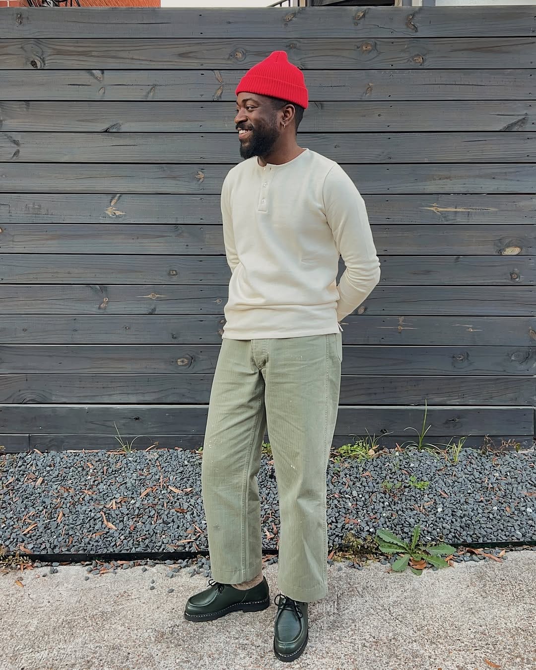 smiling man wearing a bright red beanie, cream-colored long-sleeve henley, light green pants and black lace-up leather shoes