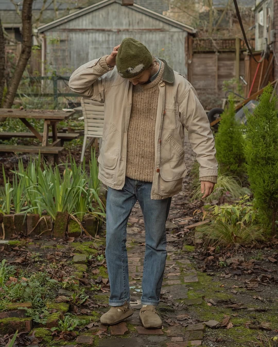 man standing outside among plants wearing a green beanie, tan chunky sweater, barn jacket, blue jeans, and birkenstock clogs