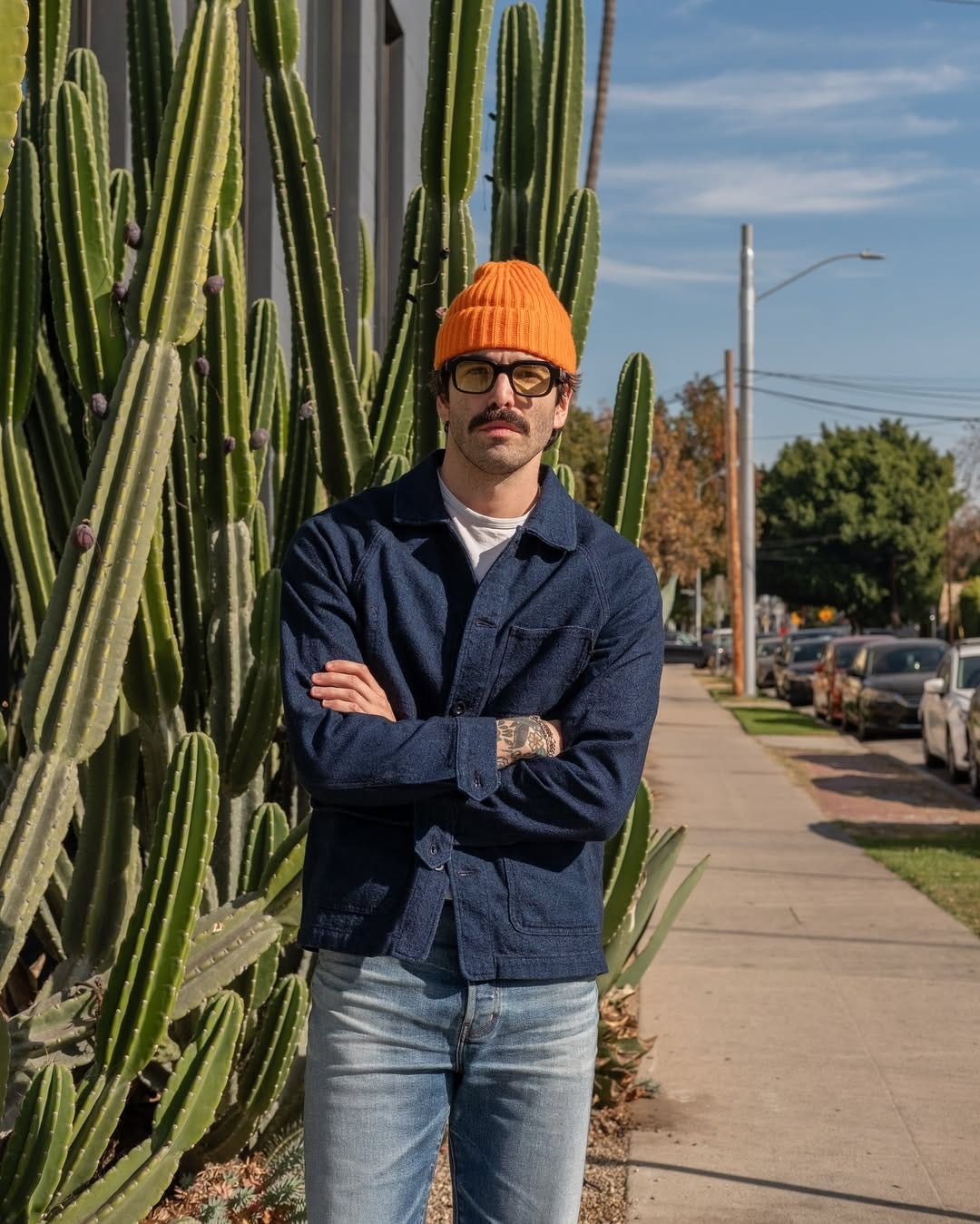 stylish man with his arms crossed wearing an orange beanie, dark frame glasses, blue chore coat, white t-shirt, and light wash jeans