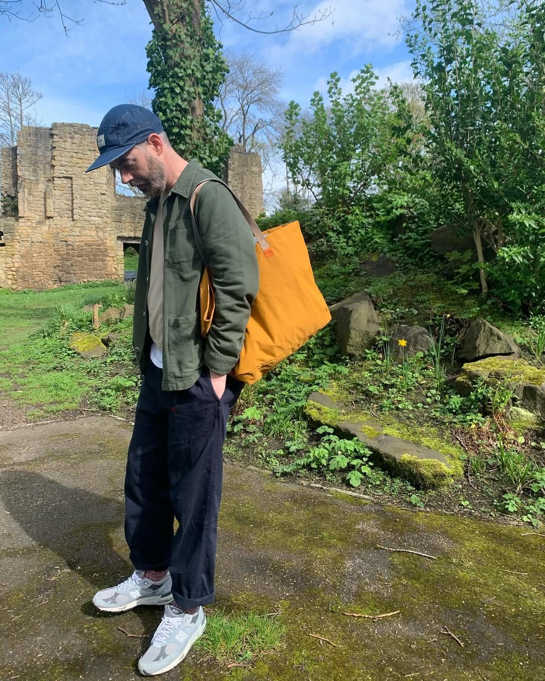 man standing outside wearing a blue 8-panel cap, green chore jacket, tan sweater, navy pants, and grey new balance sneakers, holding an orange bag