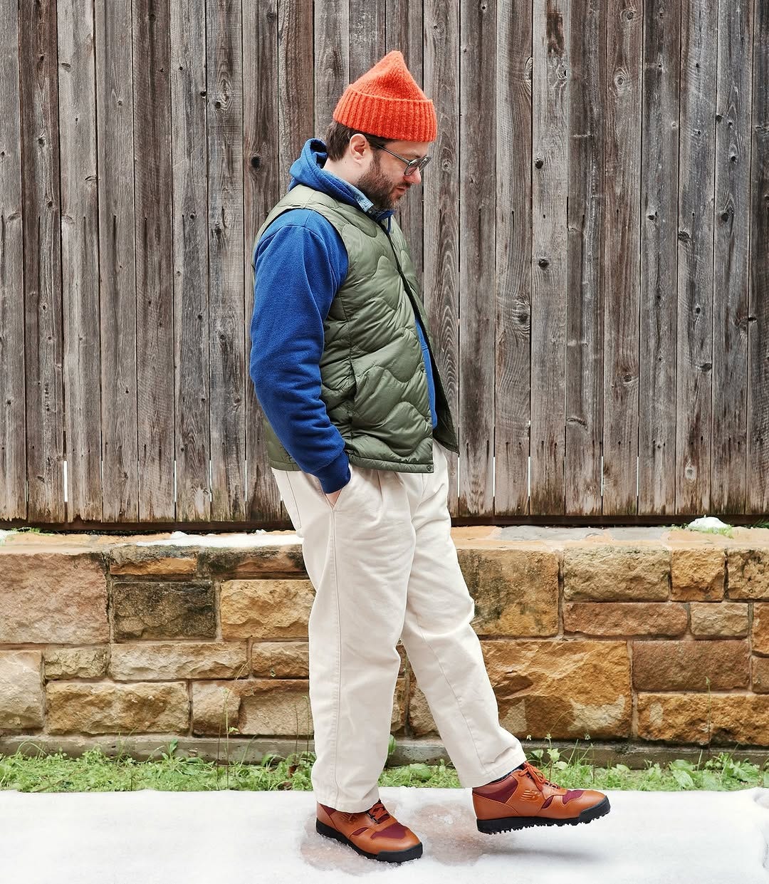 man standing outside in front of a fence wearing an orange beanie, blue hoodie sweatshirt, green quilted vest, light khakis, and brown hiking boots