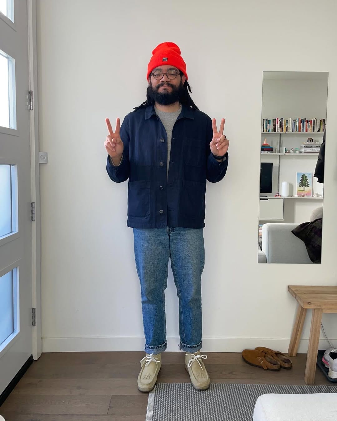 stylish man standing in a living room wearing a grey mockneck sweater under a navy chore coat with jeans and lace-up shoes making two peace signs with his hands