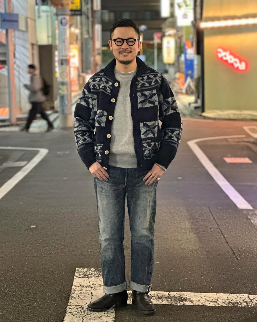 smiling man standing on a city street wearing a grey crewneck sweatshirt under a knitted patterned cardigan, dark jeans, and black boots