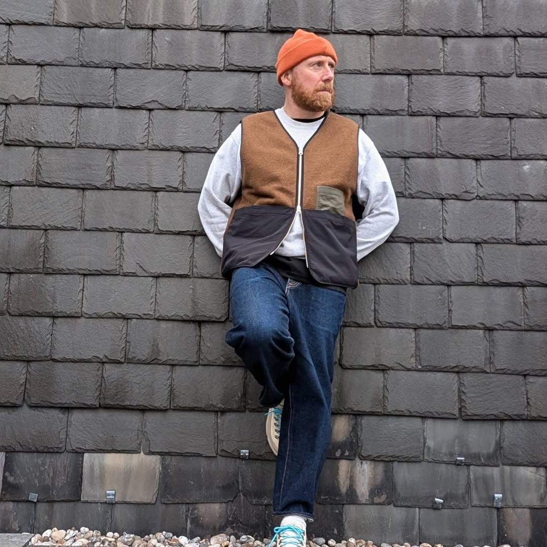 stylish man leaning against a wall wearing an orange beanie, grey long-sleeve shirt, workwear vest, blue jeans, and hiking boots