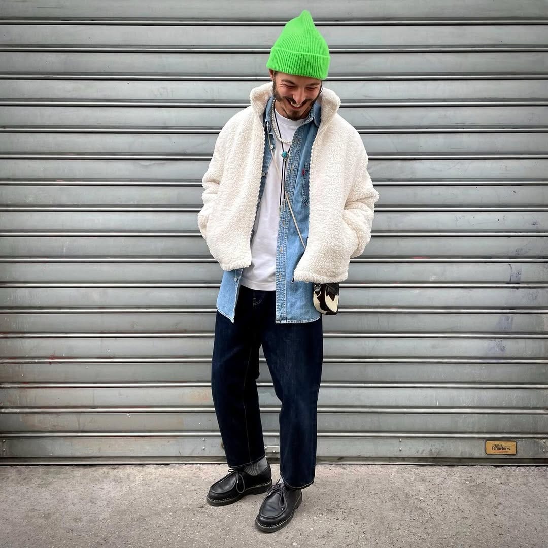 smiling man wearing a bright green beanie, fuzzy white jacket, blue denim shirt, dark pants, black leather lace-up shoes and a cross body bag