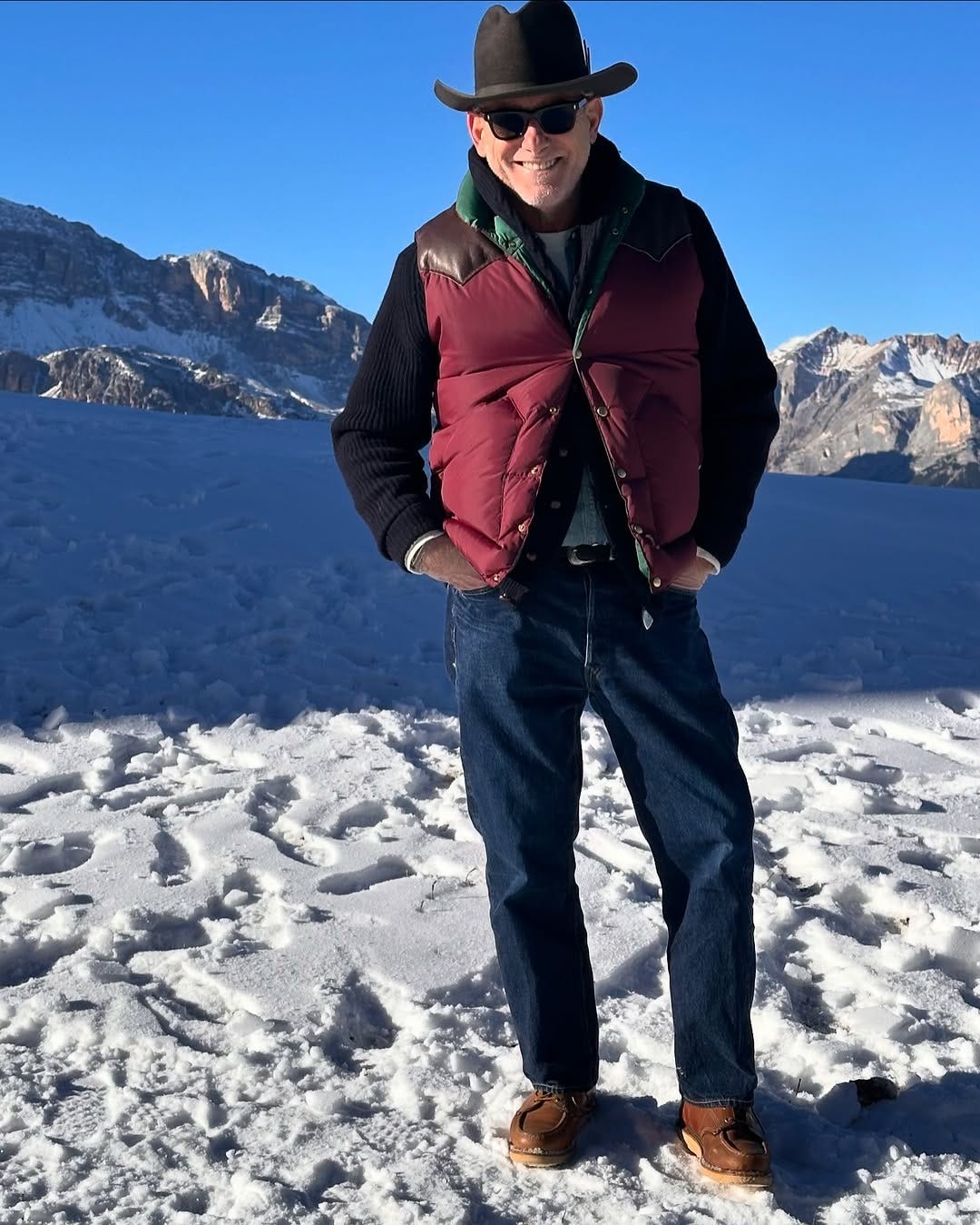 stylish older man wearing a cowboy hat, maroon puffer vest, dark sweater, and dark jeans