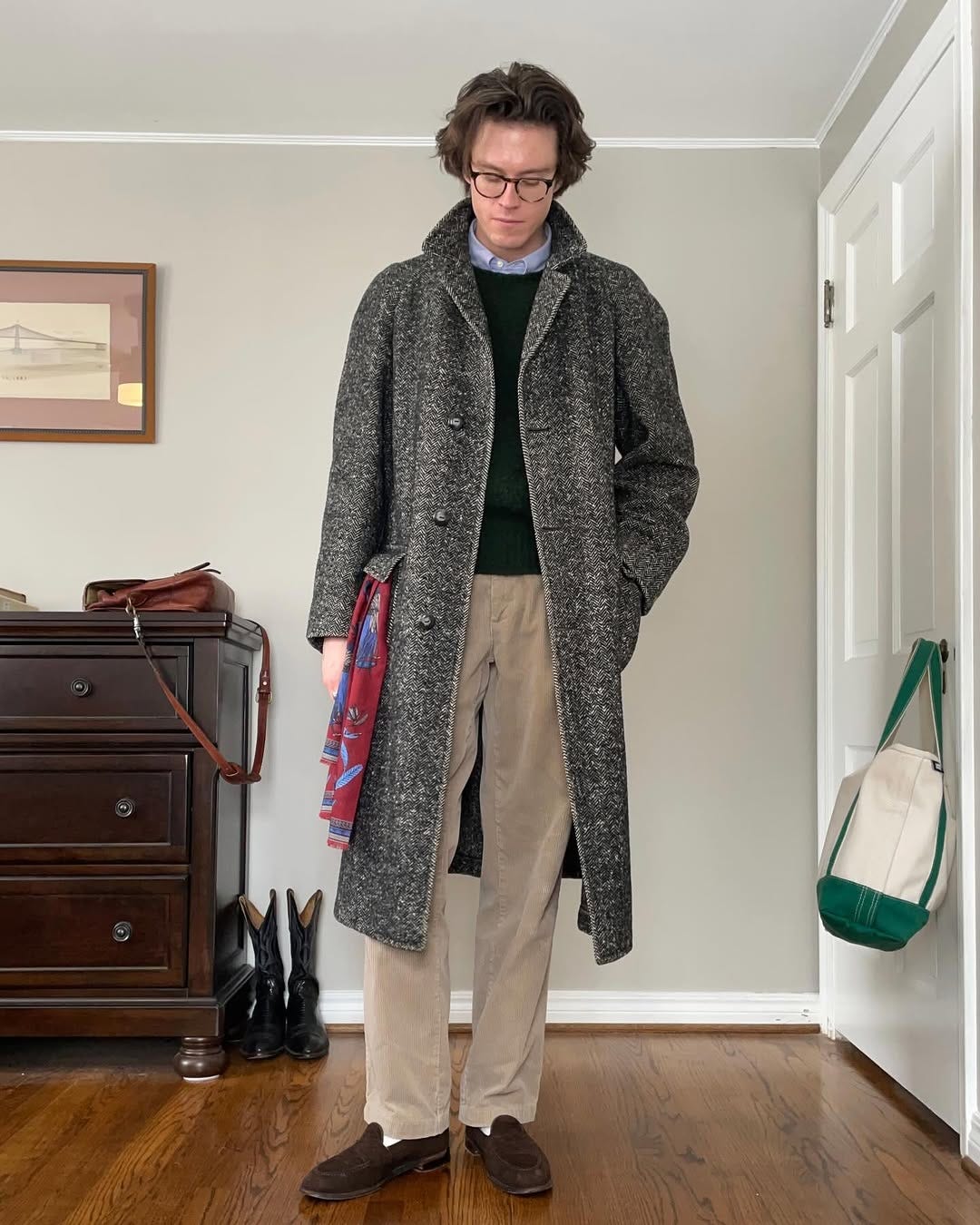 stylish man dressed in a business casual outfit of a crewneck sweater over a blue oxford shirt with tan pants and dark brown suede loafers. a patterned silk scarf is sticking out of the pocket of his patterned topcoat