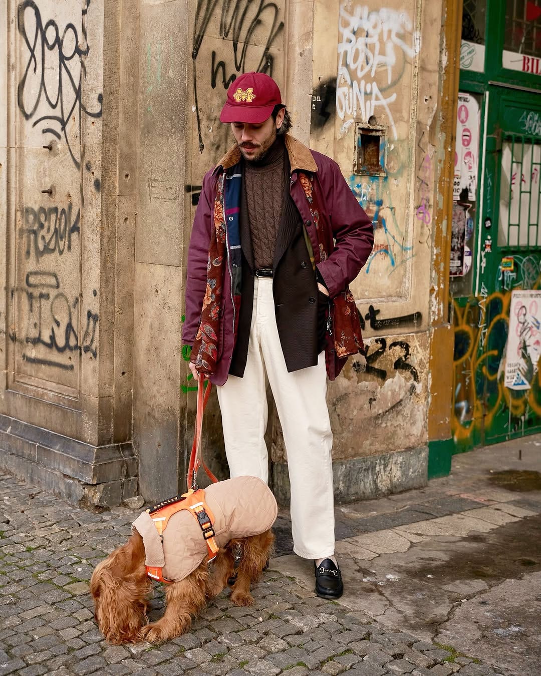 stylish man standing on a street corner with a dog, wearing a maroon jacket and patterned silk scarf