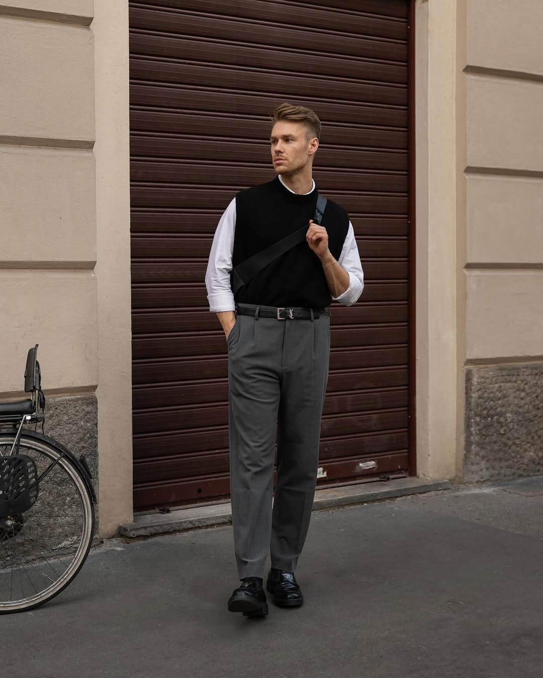 stylish man wearing a black sweater vest over a white dress shirt with grey dress pants, black leather belt, and black shoes