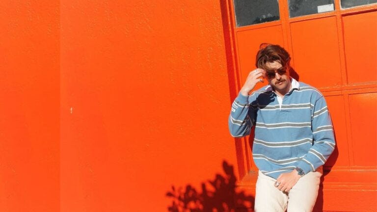 man leaning against a bright orange garage, and wearing a blue and brown striped rugby shirt with light tan chinos