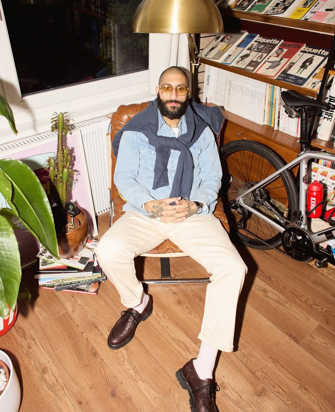 stylish man sitting on a leather chair in a leather chair wearing a blue sweatshirt around his shoulders, a light blue chambray shirt, tan pants, and brown derby shoes