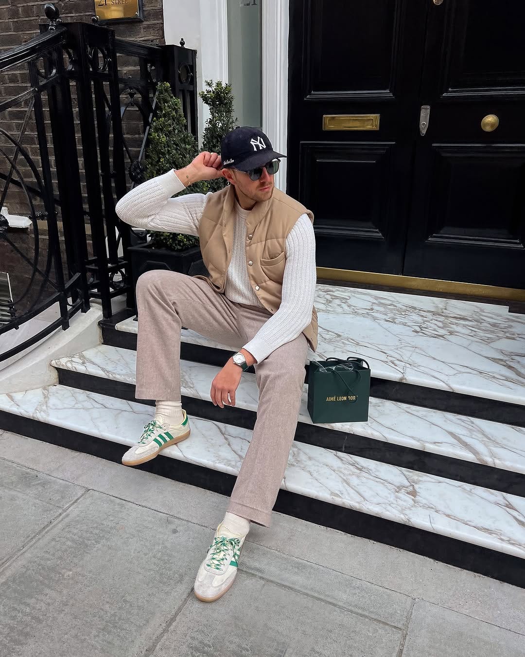 stylish man sitting on the stoop of a townhouse next to an aime leon dore shopping bag. he is wearing a navy new york yankees baseball cap, sunglasses, tan buttoned quilted vest, cream-colored sweater, tan pants, and green stripe Adidas samba sneakers