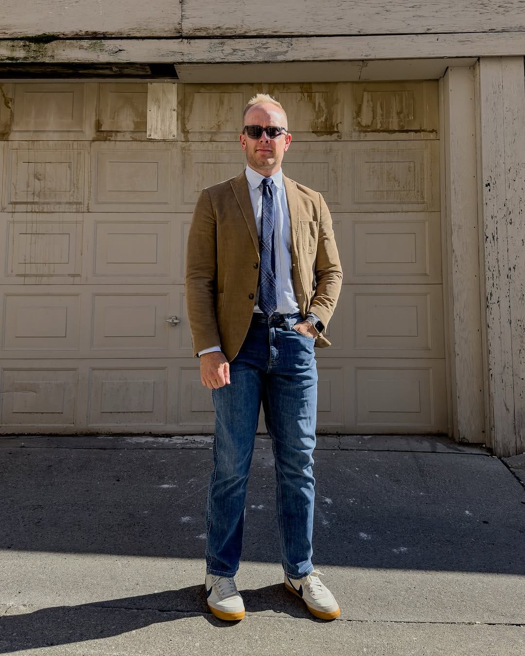 stylish man wearing a business casual outfit of a brown sport coat, dress shirt with plaid wool tie, jeans, and nike killshot sneakers