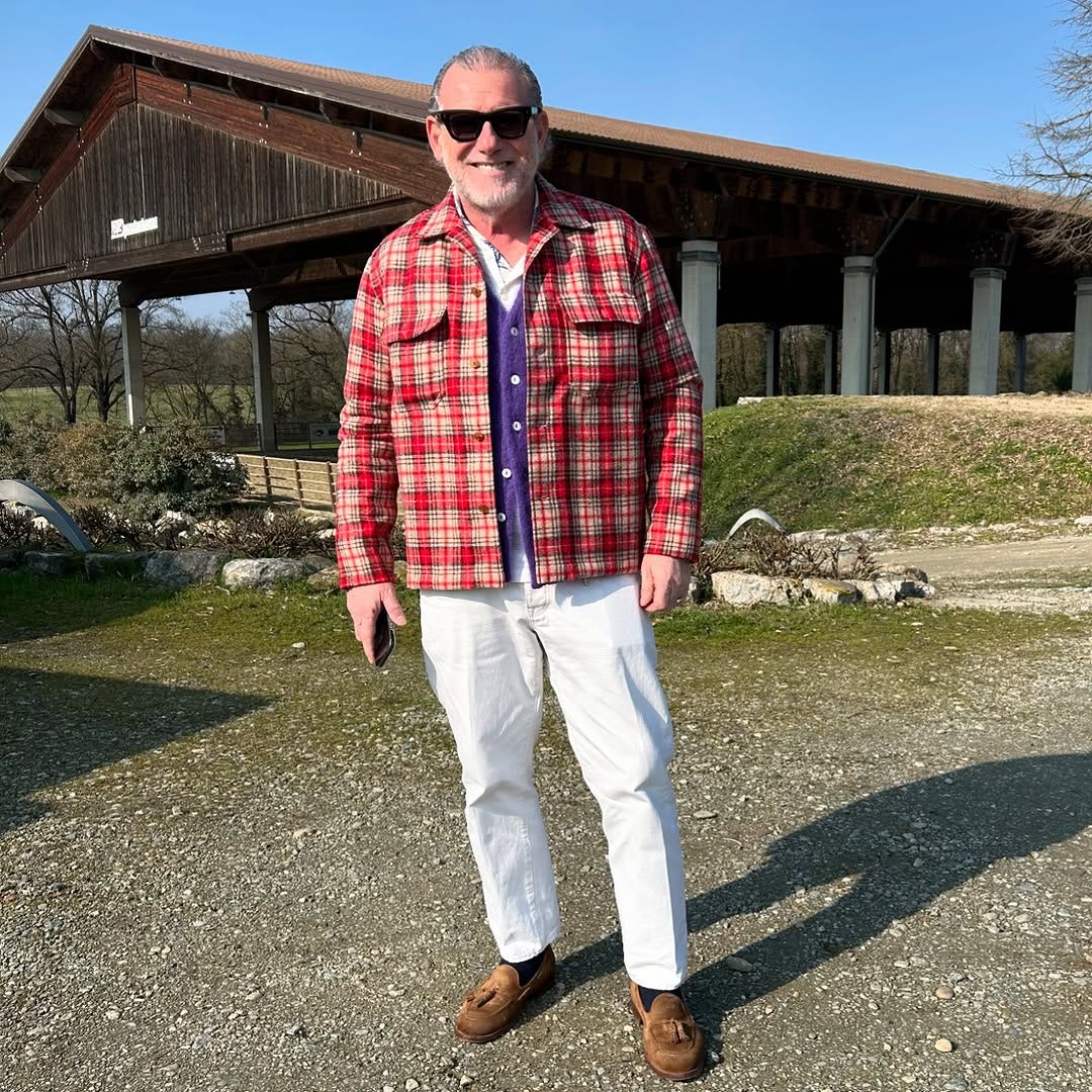 stylish older man wearing a red plaid overcoat with a purple cardigan underneath. the rest of his outfit includes white pants and brown shoes