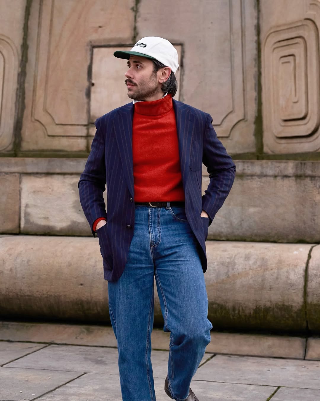 stylish man wearing a navy pinstripe blazer over a red turtleneck with a button-down shirt peeking out underneath, with jeans, a black belt, and loafers