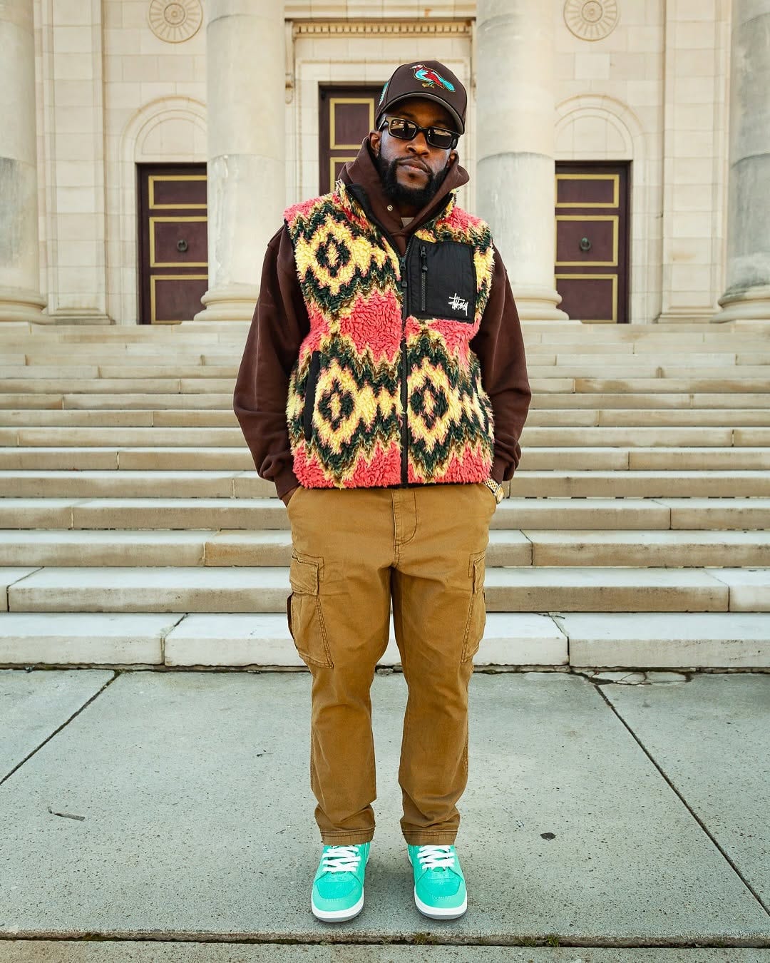 stylish man wearing a red and yellow patterned stussy fleece vest over a brown hoodie sweatshirt with tan cargo pants and aqua-colored sneakers