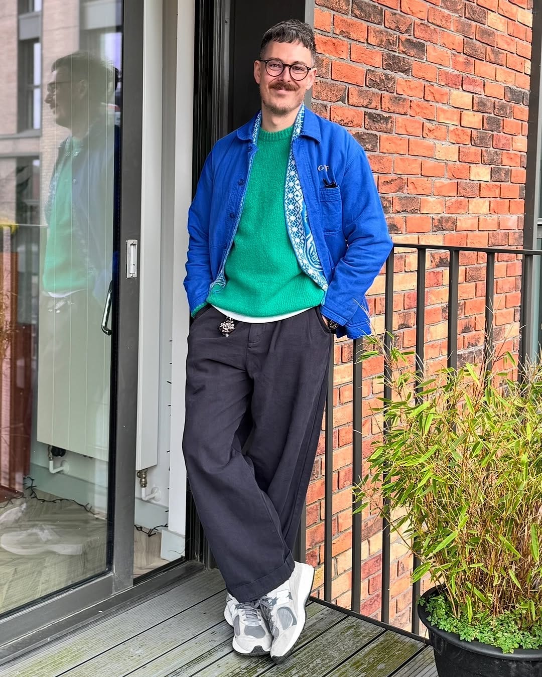 man standing on a balcony wearing a blue chore coat over a patterned blue shirt and green sweater, with wide-leg navy pants and grey sneakers