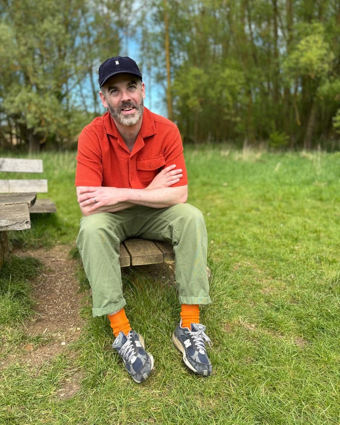 man sitting outside wearing an orange short sleeve shirt with green fatigue pants and blue new balance sneakres