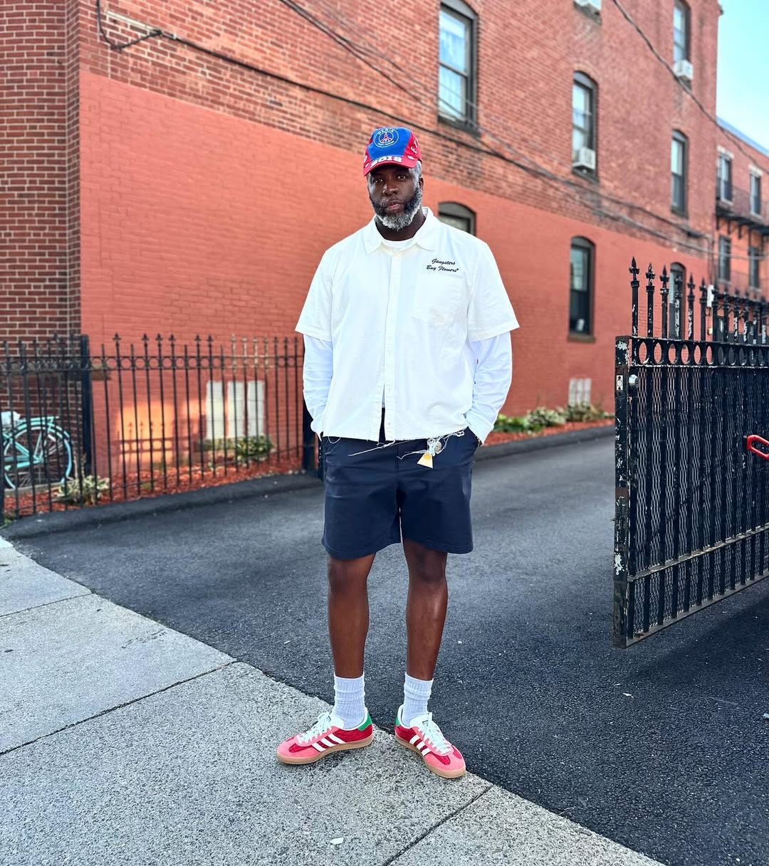 stylish. man wearing a short-sleeve shirt over a long-sleeve shirt with navy shorts and red and pink adidas samba sneakers