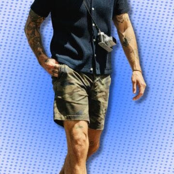 man wearing a navy sweater polo, camo shorts, and a camera across his chest