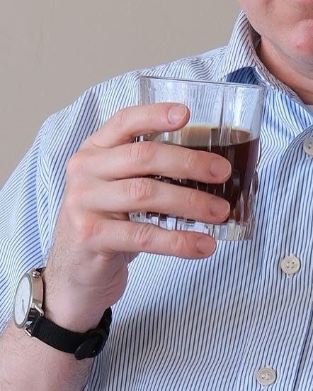 close-up of a stylish older man wearing a striped short-sleeve shirt, dark pants, and black loafers and sitting down holding a glass and reading a book