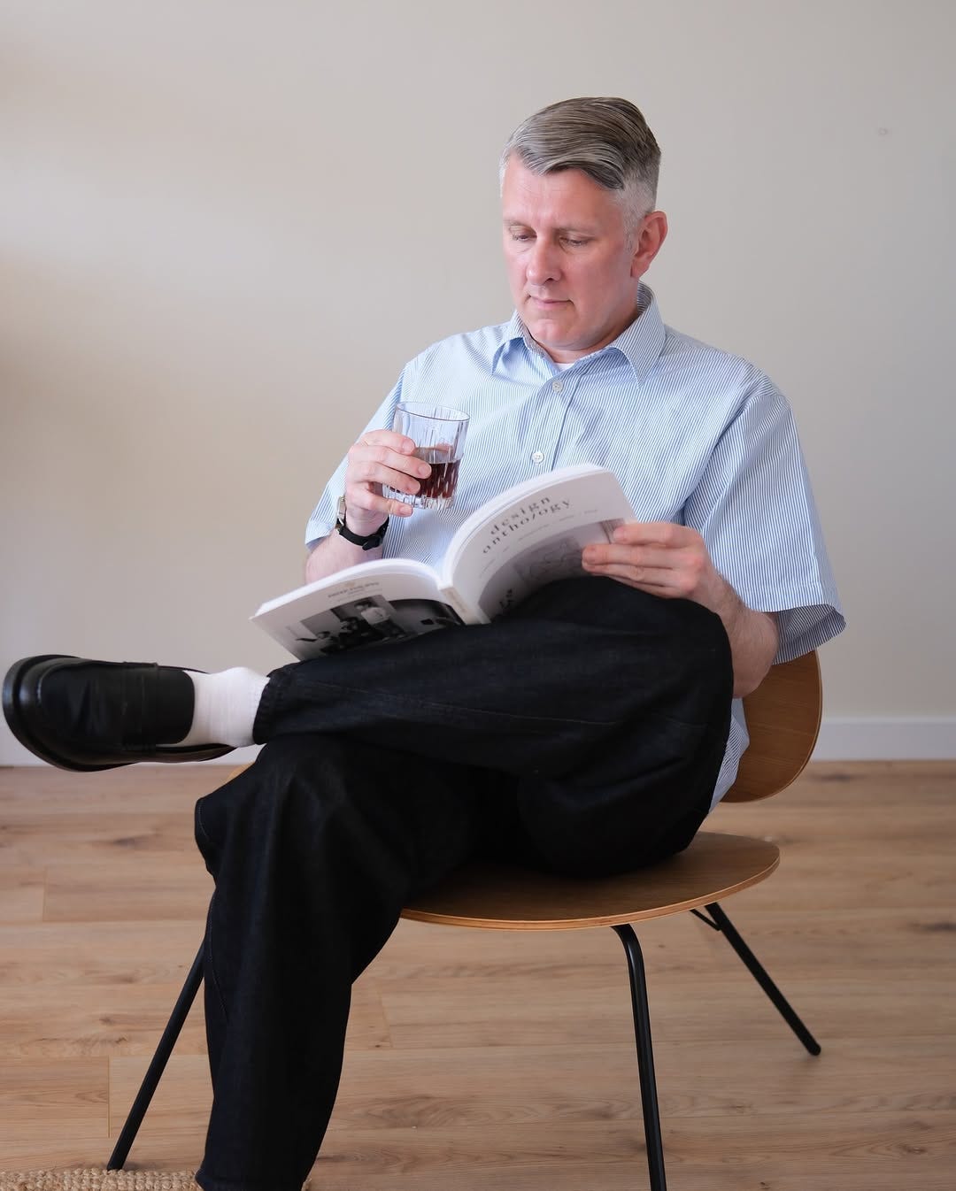 stylish older man wearing a striped short-sleeve shirt, dark pants, and black loafers and sitting down holding a glass and reading a book