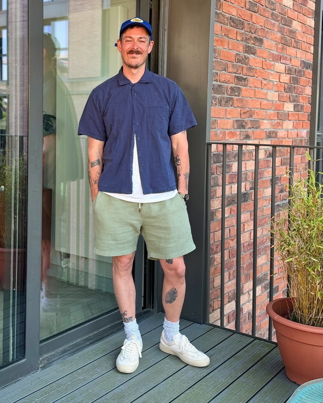 smiling man wearing a blue button-down camp shirt over a white t-shirt with green drawstring shorts and off-white sneakers and heather grey socks