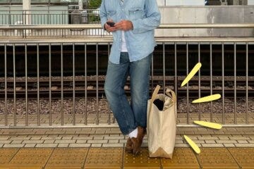 stylish man from the neck down wearing a chambray shirt, jeans, loafers, with a canvas tote bag at his feet