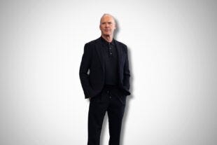 the actor Michael Keaton in a black suit with a black knit polo. his hands are in his pockets