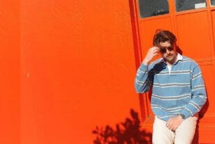 man leaning against a bright orange garage, and wearing a blue and brown striped rugby shirt with light tan chinos